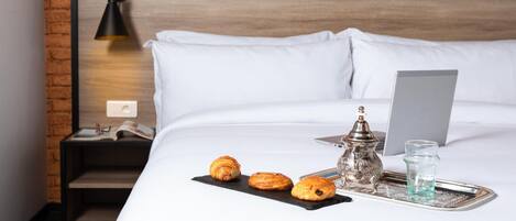 Una cama bien hecha con ropa blanca, una computadora portátil, una bandeja con pasteles, un hervidor de agua y un vaso de agua en una habitación de hotel.
