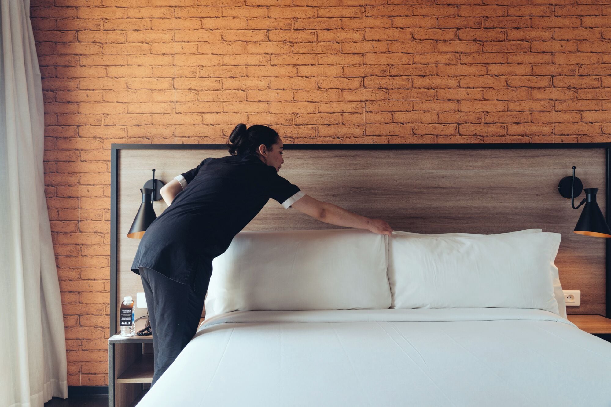 Una habitación de hotel con pared de ladrillo, una cama con ropa de cama blanca, una mesita de noche con lámpara y una botella de agua sobre la mesita.
