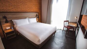 A hotel room with a brick wall, a wooden bed, a nightstand with a lamp, a chair, and a desk with a lamp.