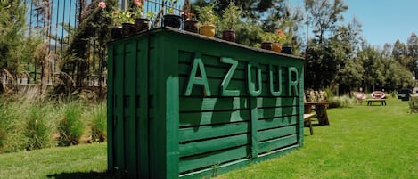 Una estructura de madera verde con el letrero "AZOUER" y plantas en macetas en la parte superior, rodeada de césped y árboles.