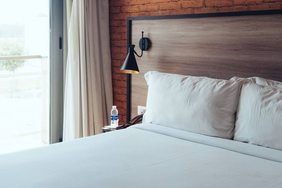 Une chambre d’hôtel avec un mur en briques, un lit avec du linge de maison blanc, une lampe noire, une bouteille d’eau et un téléphone sur une table de chevet.