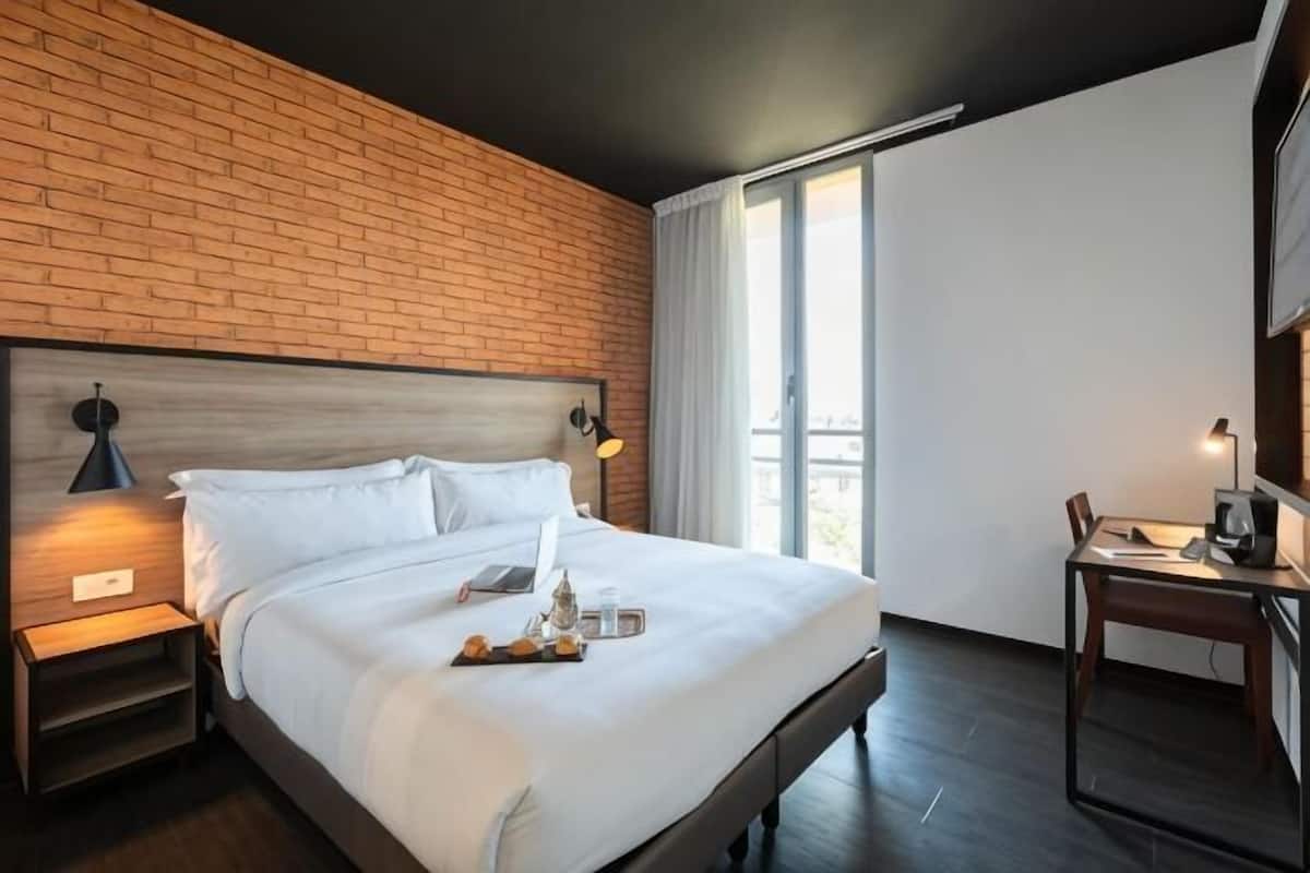 Une chambre d’hôtel avec un mur en briques, un grand lit avec du linge de maison blanc, un bureau avec une chaise et une fenêtre avec des rideaux.