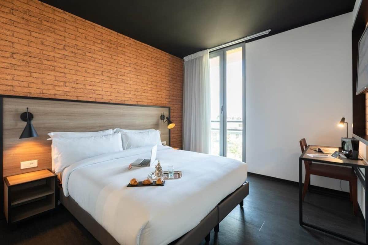 Une chambre d’hôtel moderne avec un mur en briques, un grand lit avec du linge de lit blanc, un bureau avec un ordinateur et une vue sur la ville par la fenêtre.