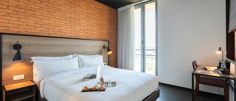 Una habitación de hotel moderna con pared de ladrillo, una cama grande con ropa blanca, un escritorio con una computadora y vista a la ciudad a través de la ventana.