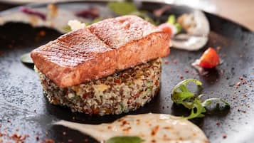 A dish with a crispy top, possibly a burger or sandwich, on a dark plate with a side of greens and a creamy sauce.