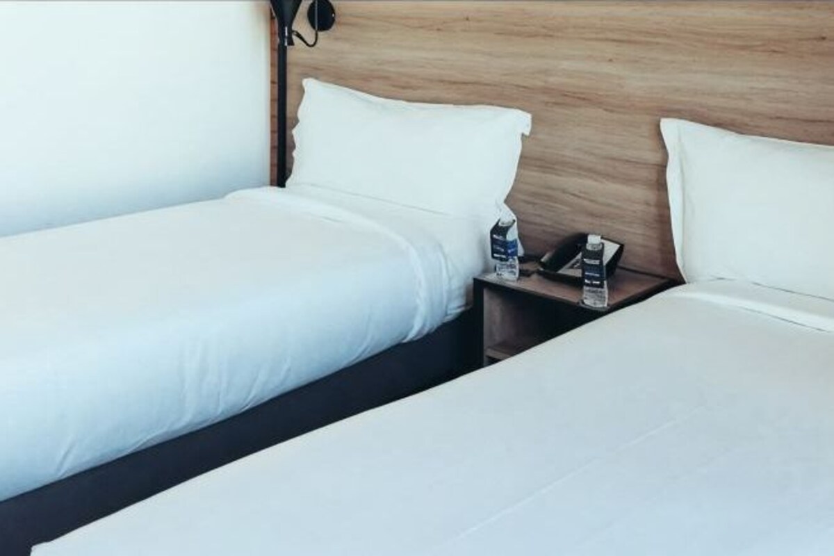 Una habitación de hotel con dos camas individuales, un cabecero de madera, una mesita de noche con un teléfono y una botella, y una lámpara de pared.