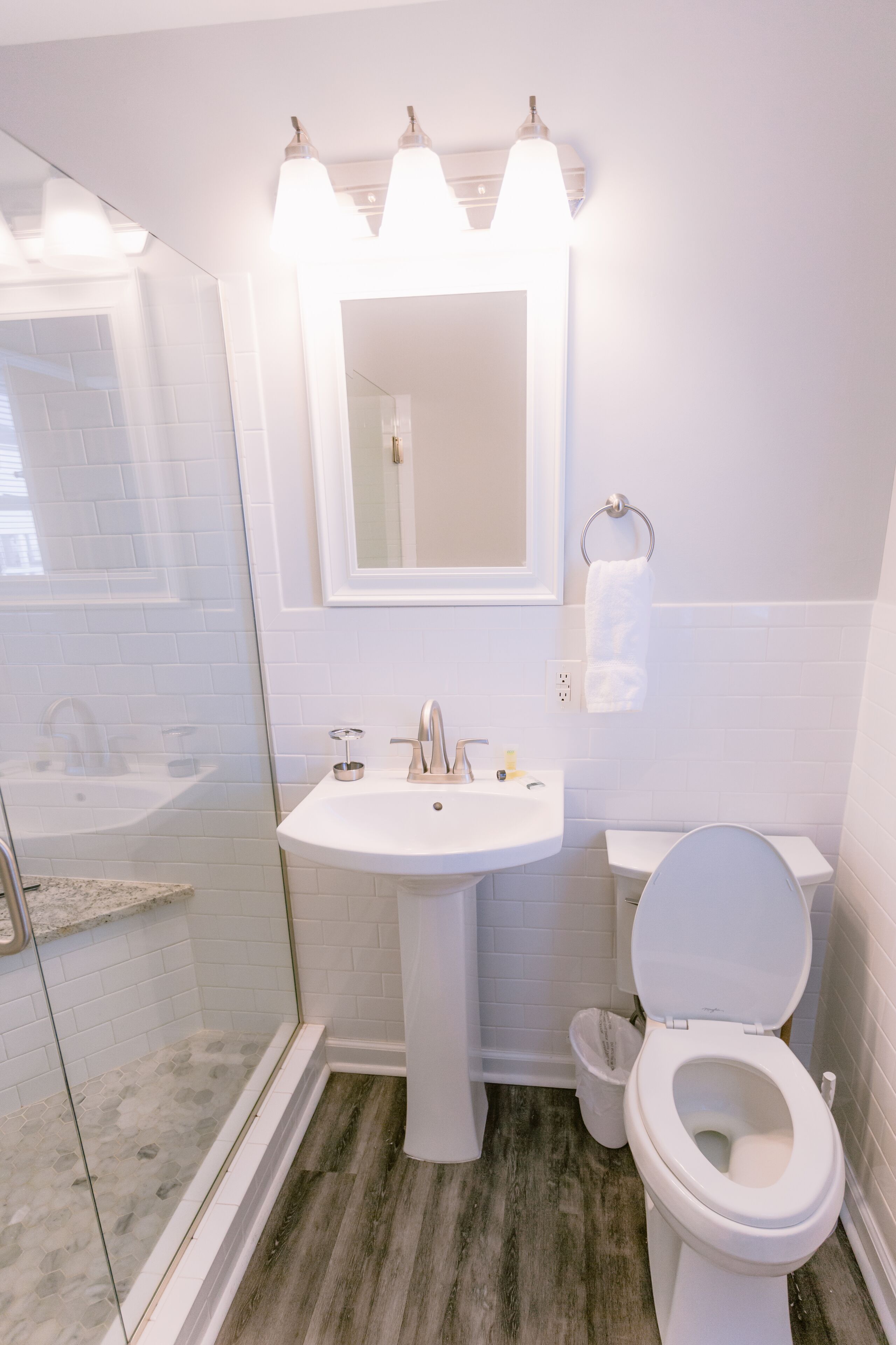 Un baño con lavamanos blanco, espejo, inodoro, cabina de ducha de vidrio y suelo de madera.