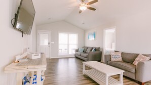 A spacious living room with a high ceiling, a ceiling fan, a flat-screen TV mounted on the wall, a gray sofa with decorative pillows, a white coffee table, and a wooden floor. - 3 bedroom-2 bathroom Beach house in the heart of Garden City/Murrells Inlet (Murrells Inlet)