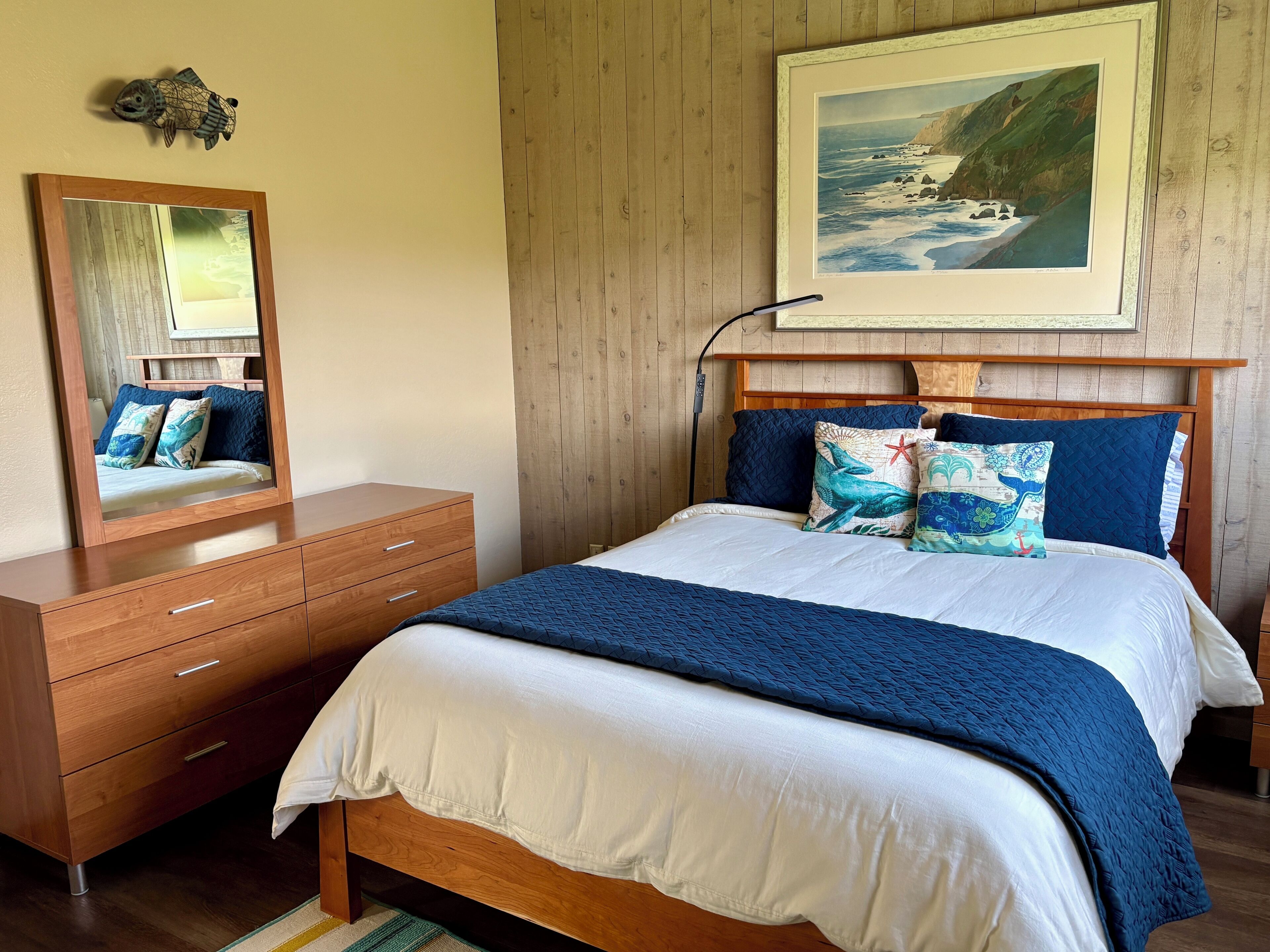 A bedroom with a wooden bed, white bedding, and a painting of a coastal scene.