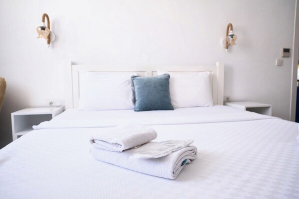 Una cama bien hecha con una toalla blanca doblada y una almohada azul, mesitas de noche blancas e lámparas fijadas a la pared.