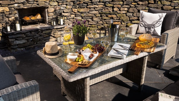 An outdoor dining area with wicker furniture, a stone wall, a fireplace, and a table set with food and drinks.