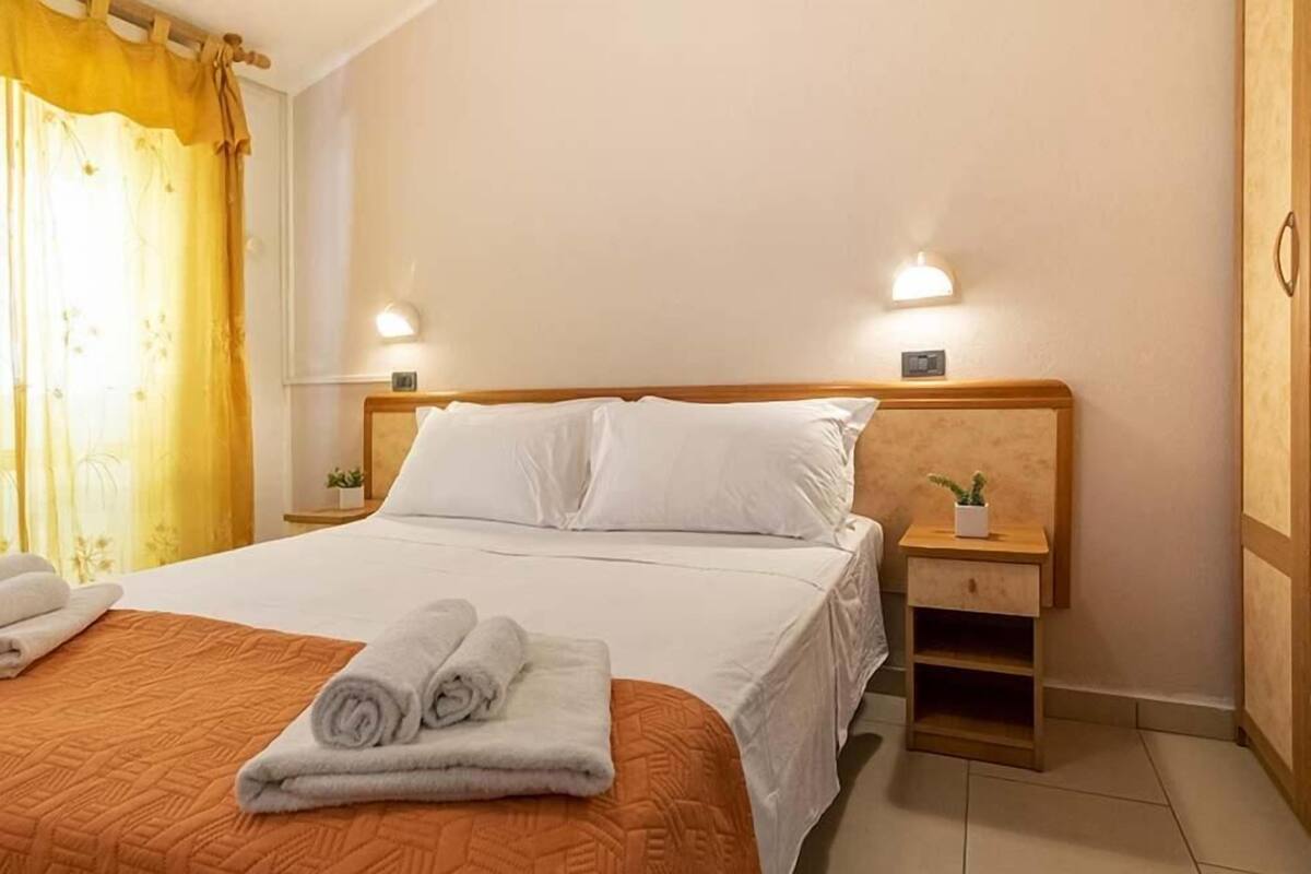 A neatly made bed with white linens and rolled towels, a wooden nightstand with a lamp, and a small potted plant.