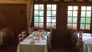 Un comedor con mesas y sillas de madera, suelo con patrón de espiga y grandes ventanales con vistas al verdor.