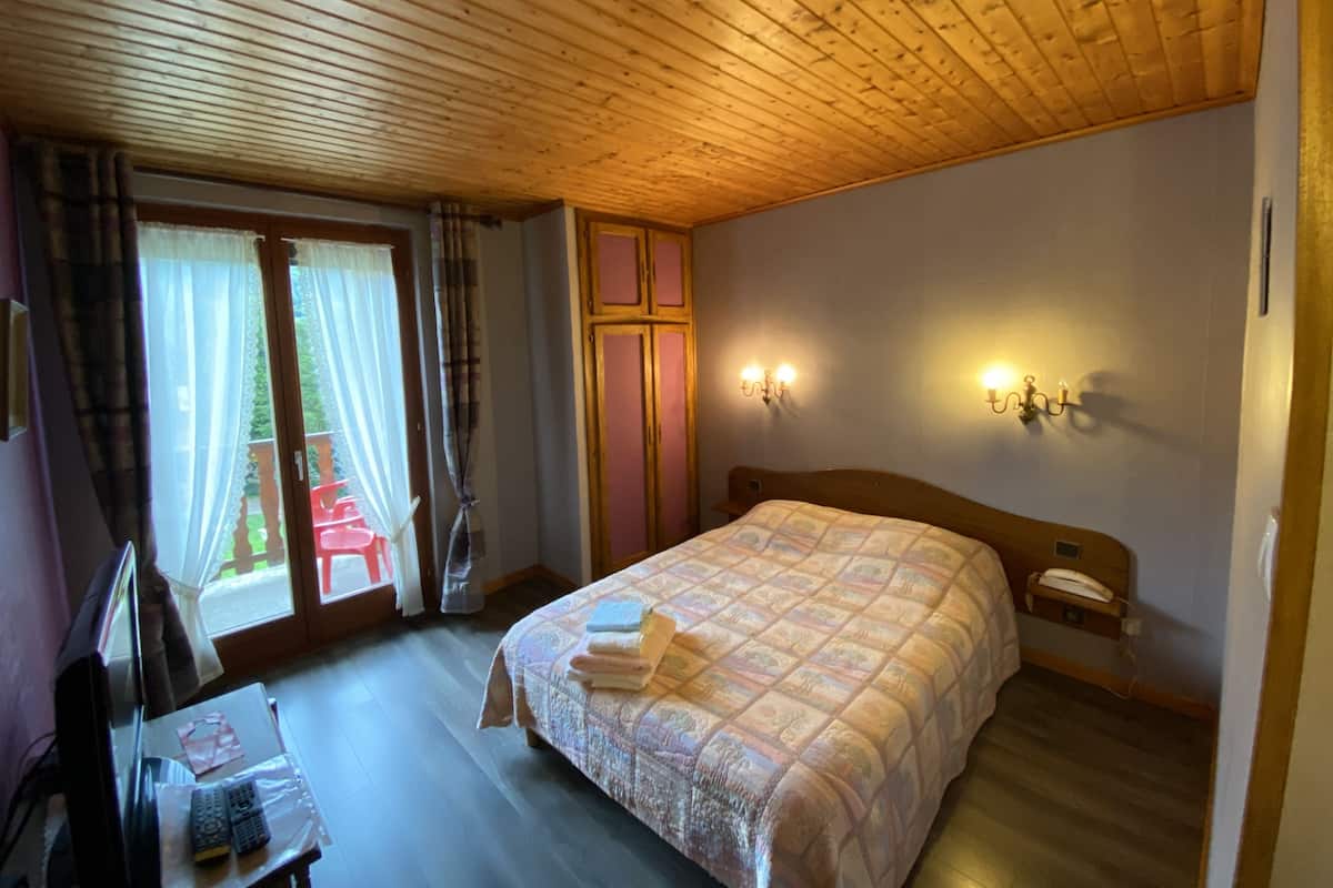 A bedroom with a wooden ceiling, a bed with a quilt, a nightstand with a lamp, a window with curtains, and a view of a balcony with red chairs.
