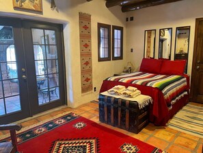 Ein Zimmer mit einem Bett, einer Kommode, einer Tür mit Glasfeldern, einem wandmontierten Spiegel und einem gemusterten Teppich.