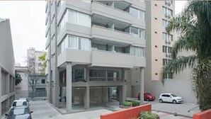 A modern multi-story building with a flat facade, large windows, and a red accent wall. There are cars parked on the street in front. - HOMETANGO ABASTO (CABA)