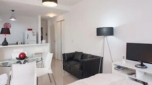 A modern, minimalist room with a bed, a black leather sofa, a dining table with chairs, a flat-screen TV, and a floor lamp. - HOMETANGO ABASTO (CABA)