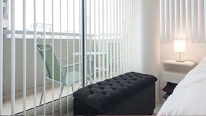 A hotel room with a black ottoman, a small bedside table with a lamp, and a view through a window with vertical blinds. - HOMETANGO ABASTO (CABA)