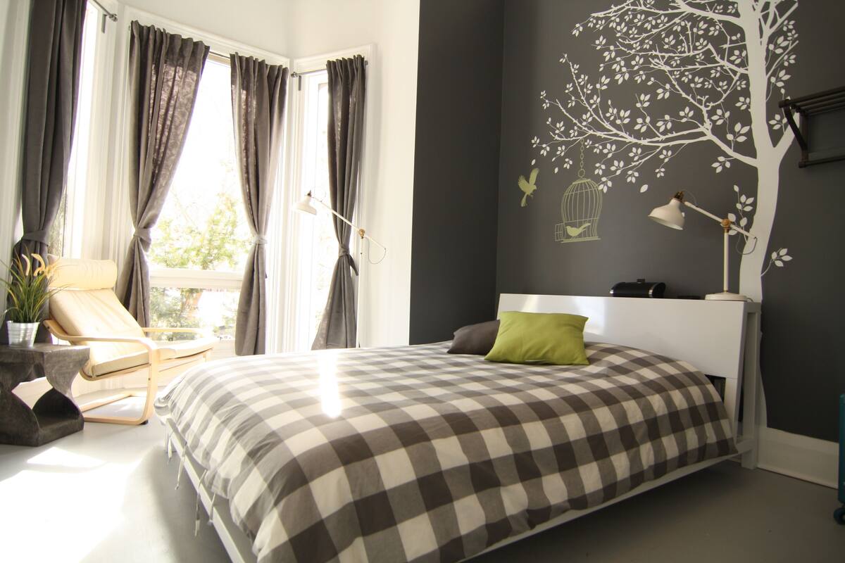 Een slaapkamer met een geruit beddegoed, een groen kussen, een bedlampje en een muurschildering van een boom met vogels.