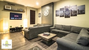 Un salon moderne avec un canapé gris, une table basse sur laquelle sont posés des livres, une horloge murale, un téléviseur diffusant Netflix et des tableaux encadrés représentant des paysages urbains.