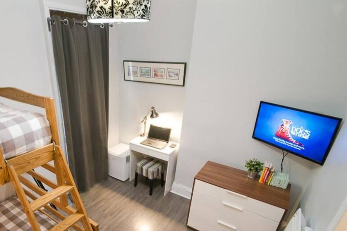 Een slaapkamer met een houten stapelbed, een bureau met een laptop, een televisie en een kleine plant.