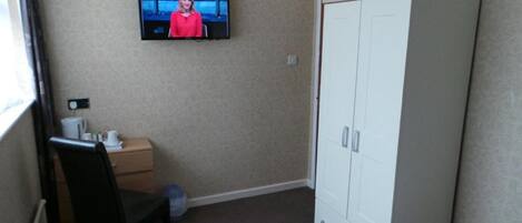 A room with a television mounted on the wall displaying a news broadcast, a white wardrobe, a small wooden nightstand with a cup and a tissue box, and a black chair.