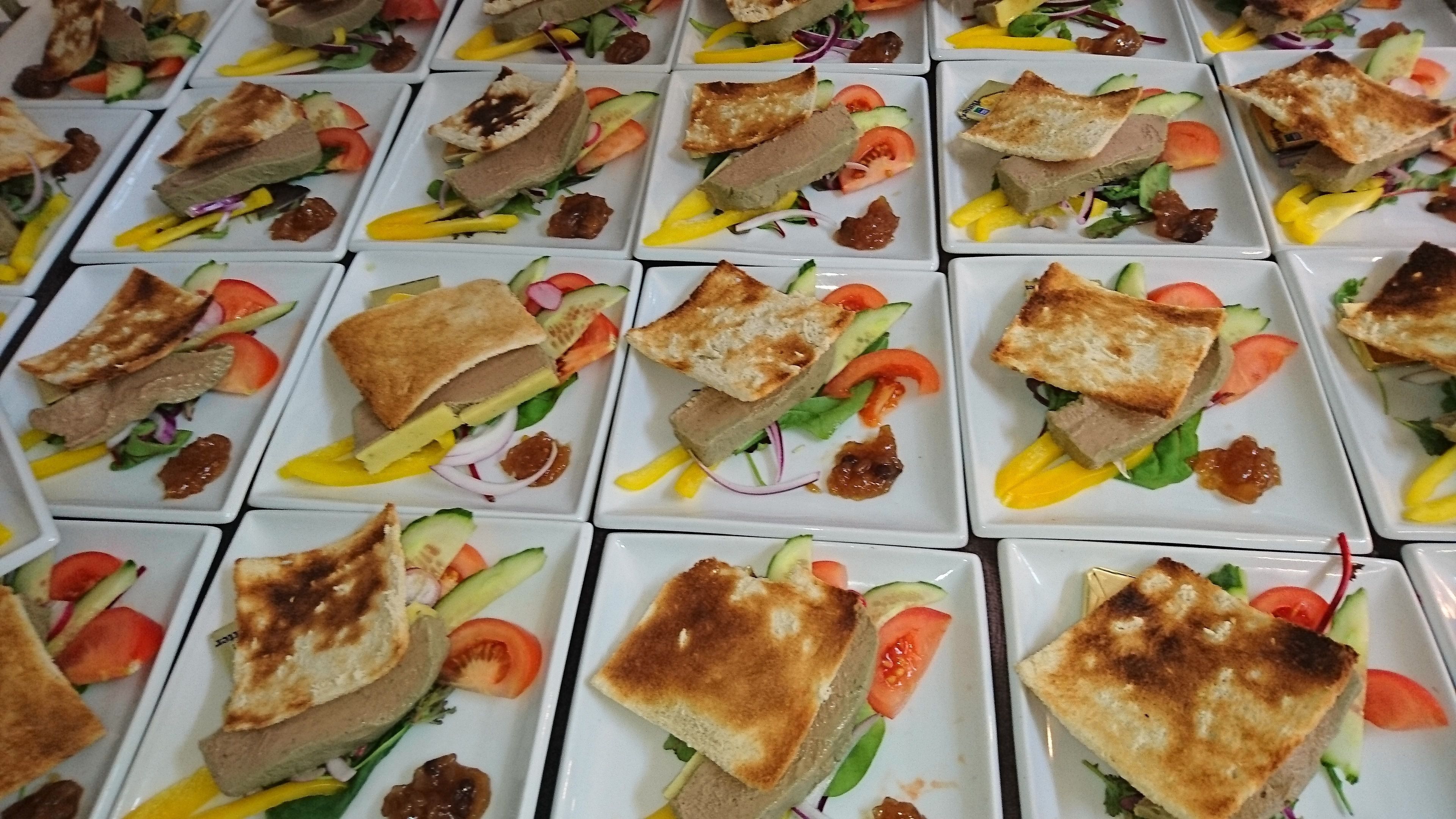 A table with multiple square plates of food, each containing sandwiches, vegetables, and condiments.