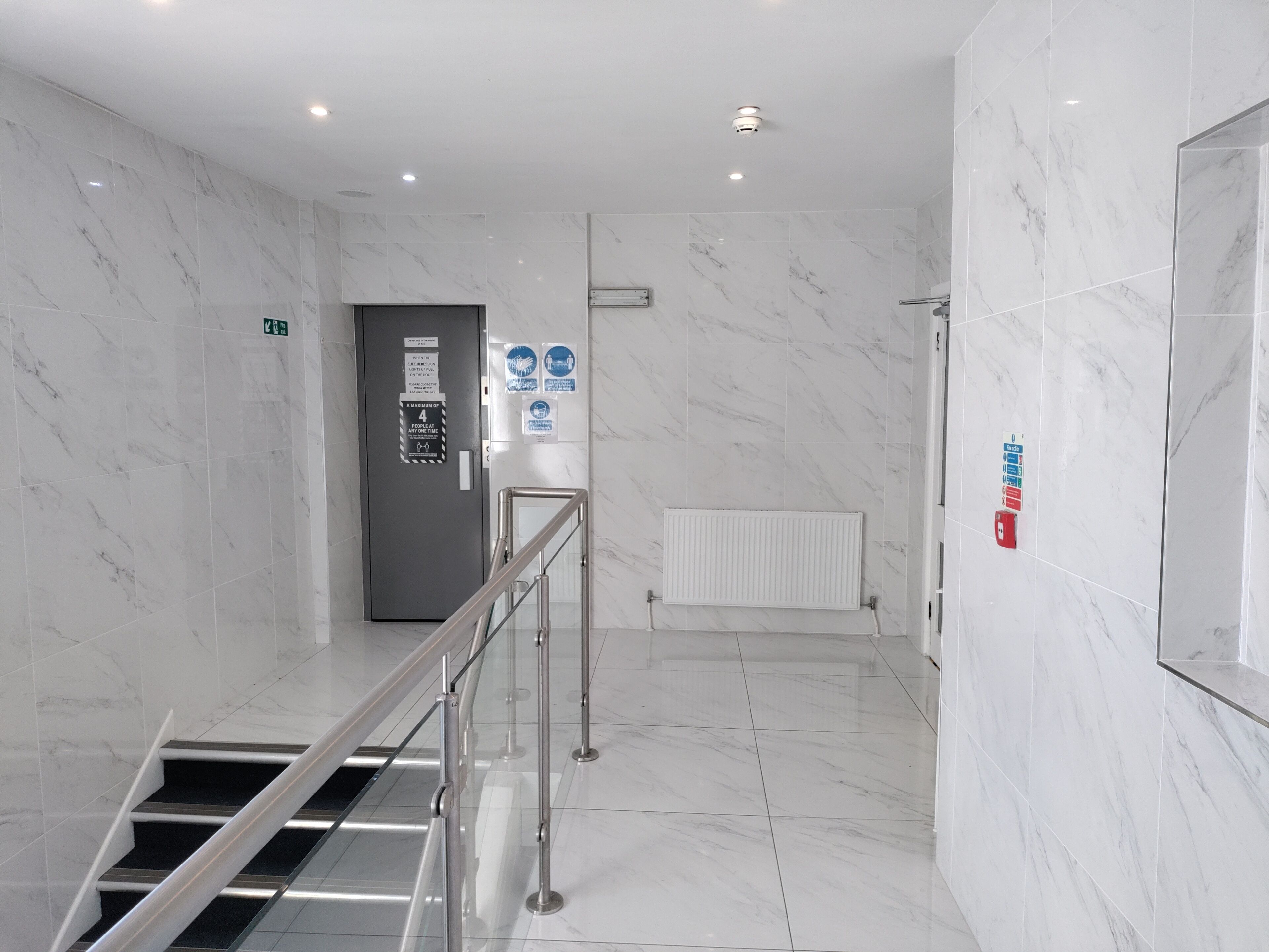 A modern hotel lobby with marble floors, a staircase, a reception desk, and a security camera.