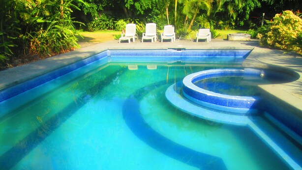 Una piscina con un jacuzzi circular, rodeado de sillones blancos y abundante vegetación.