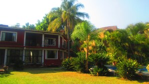 Un edificio de dos pisos con fachada roja y detalles blancos, rodeado de abundante follaje y palmeras.
