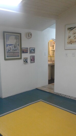 Un couloir avec un sol bleu et jaune, une affiche encadrée représentant une course automobile, une horloge et, au fond, un coin lavabo.