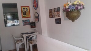 Une pièce avec un mur blanc orné d’anciennes affiches sur le thème de l’automobile et de la moto, une petite table blanche avec une chaise, et une composition florale suspendue.