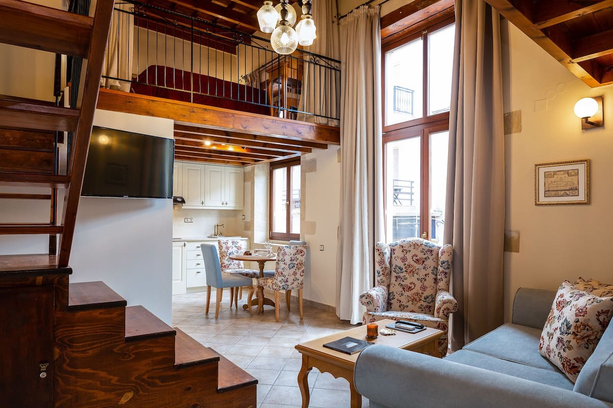 A spacious living area with a high ceiling, a staircase, a dining area, and large windows.