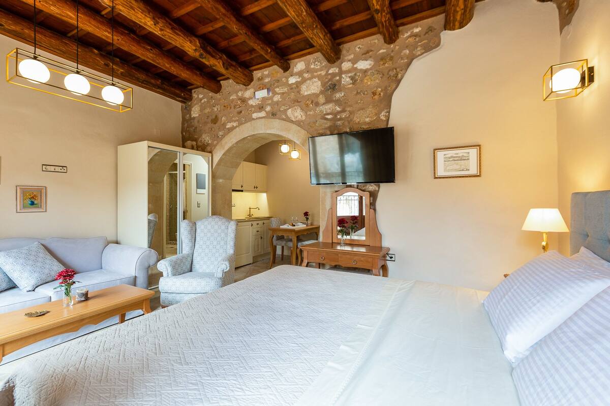 A bedroom with a large bed, a wooden desk, a chair, a sofa, and a TV mounted on the wall.