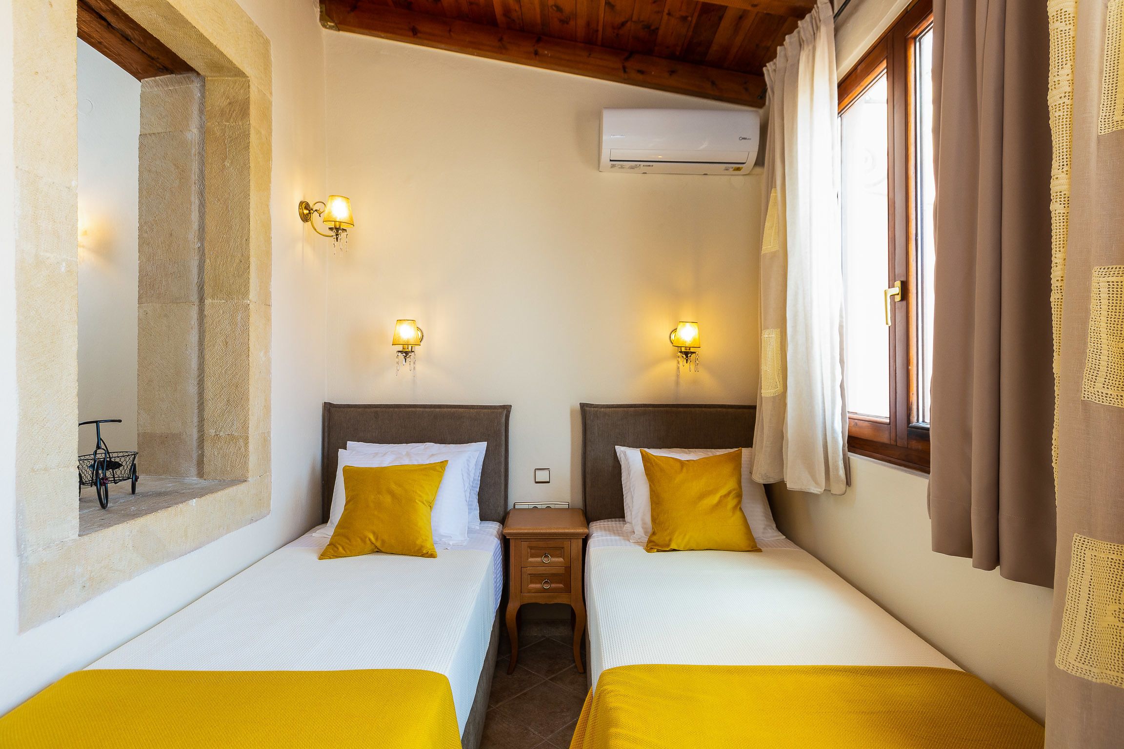 Ein Hotelzimmer mit zwei Betten, jeweils mit Kopfstütze, einem kleinen Nachttisch und einer wandmontierten Klimaanlage.