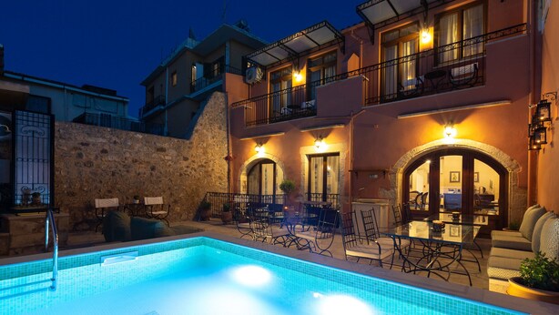 A swimming pool with a stone wall and outdoor furniture, surrounded by a building with balconies and warm lighting. - Phaedra Suites (Rethymno)