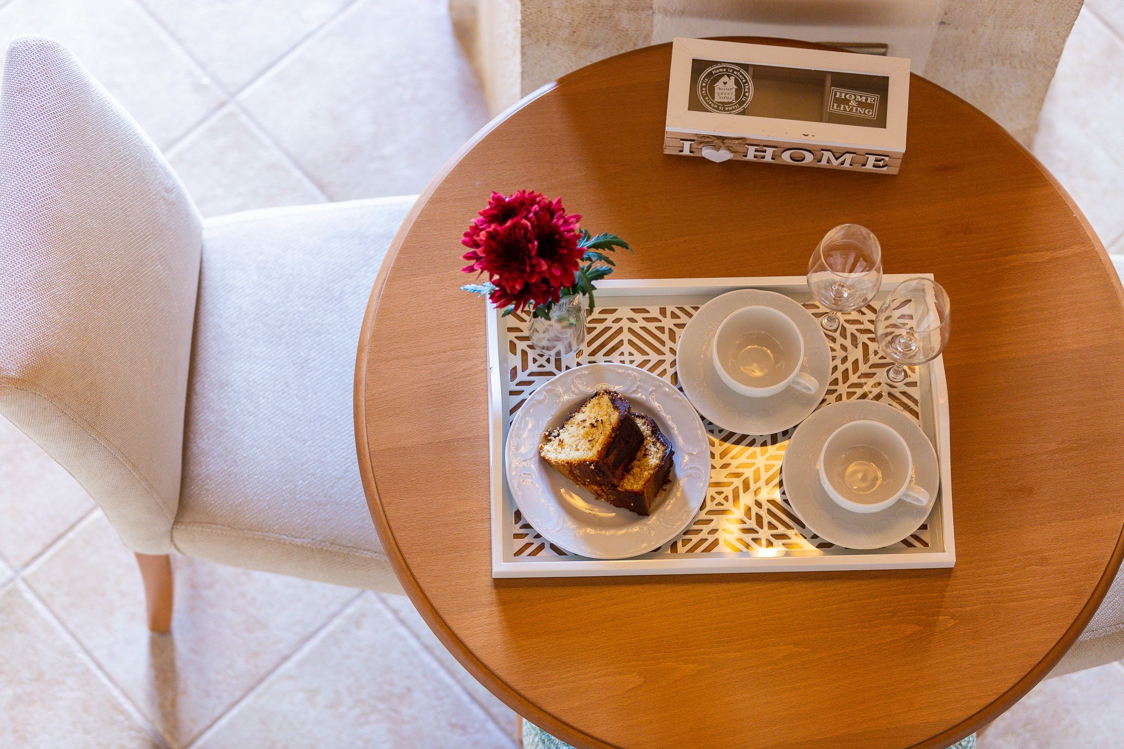 A round wooden table with a tray containing a slice of cake, two white cups, and a glass of water, accompanied by a vase with red flowers.