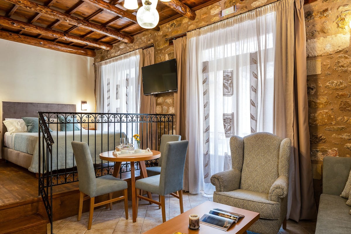 A cozy room with a bed, a dining table with chairs, a TV mounted on the wall, and large windows with curtains.