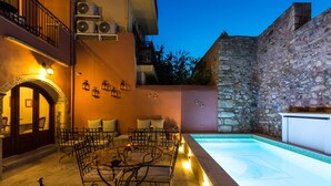 An outdoor pool area with a stone wall, a seating area with chairs and tables, and a pool with a tiled edge. - Phaedra Suites (Rethymno)