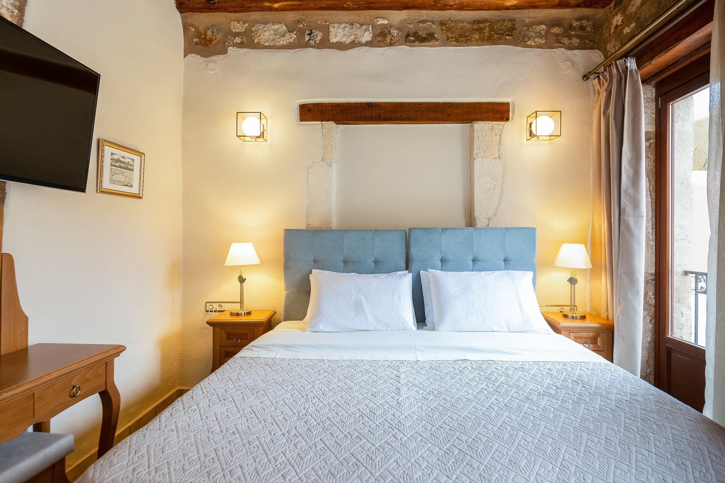 A bedroom with a bed, two bedside tables with lamps, a wooden headboard, and a television on the wall.