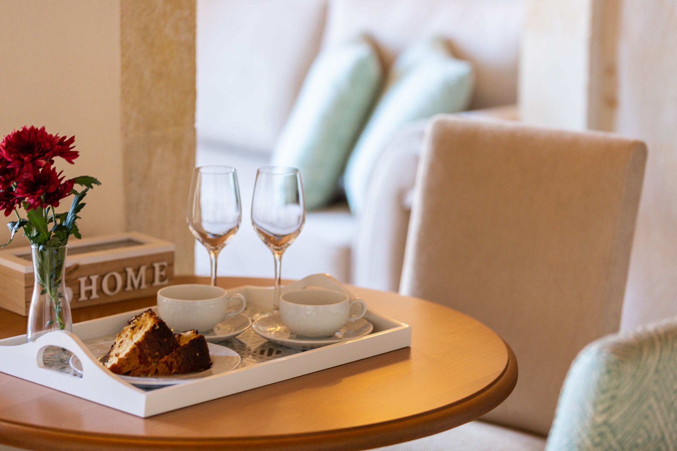 Un plateau avec une part de gâteau, deux tasses blanches et deux verres à vin, posés sur une table ronde en bois.