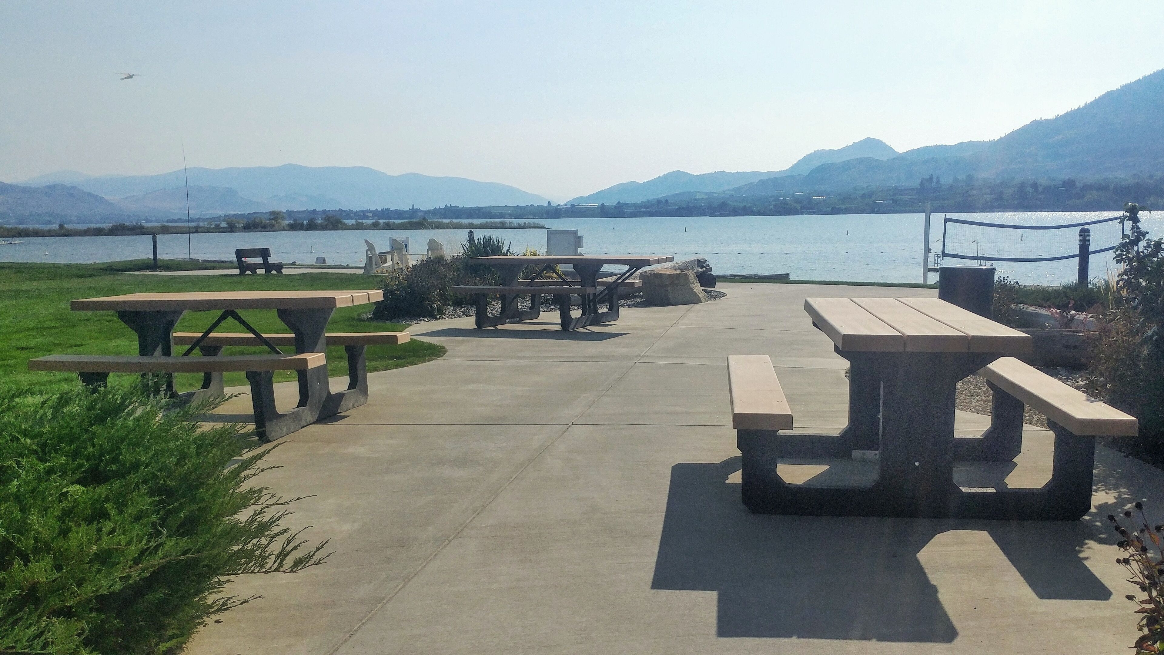A lakeside picnic area with concrete benches and picnic tables, surrounded by greenery and mountains in the background.