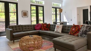 Una sala de estar moderna con un sofá modular, una mesa de centro de mimbre redonda y ventanas amplias con vista a un jardín.