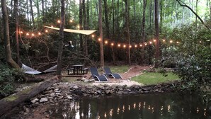 Un área boscosa y tranquila con un pequeño estanque, luces colgantes, una hamaca y una mesa de picnic.