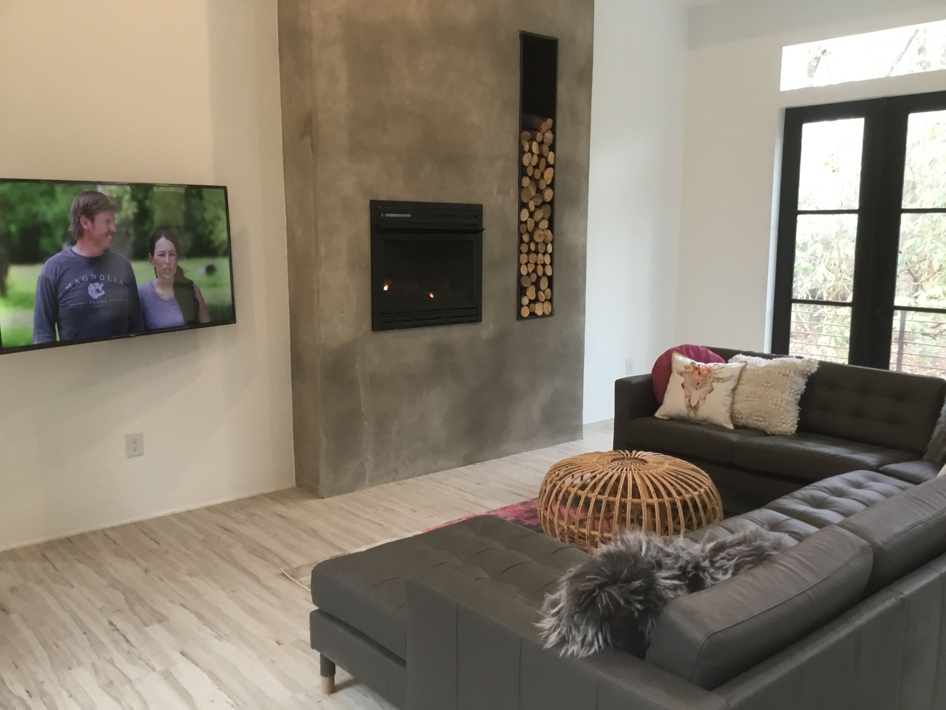 A modern living room with a grey sofa, a television mounted on the wall, a fireplace, and a large window.