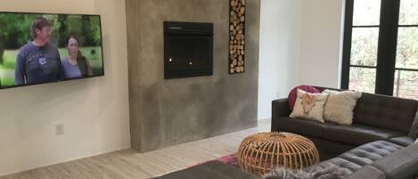A modern living room with a grey sofa, a television mounted on the wall, a fireplace, and a large window.