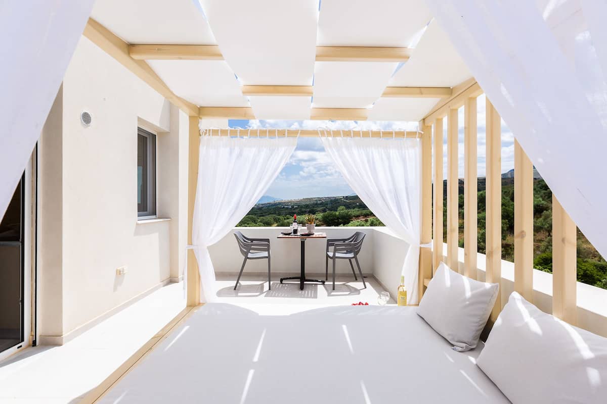 Ein Balkon mit Tisch und Stühlen, ein Bett mit Kissen und Blick auf Grünflächen und Hügel.