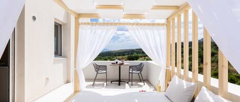 A balcony with a table and chairs, a bed with pillows, and a view of greenery and hills.