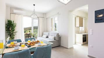 Ein stilvoller Essbereich mit einem Glastisch, blauen Stühlen und Frühstücksangeboten. Ein graues Sofa und ein großes Fenster bieten Ausblick ins Freie.