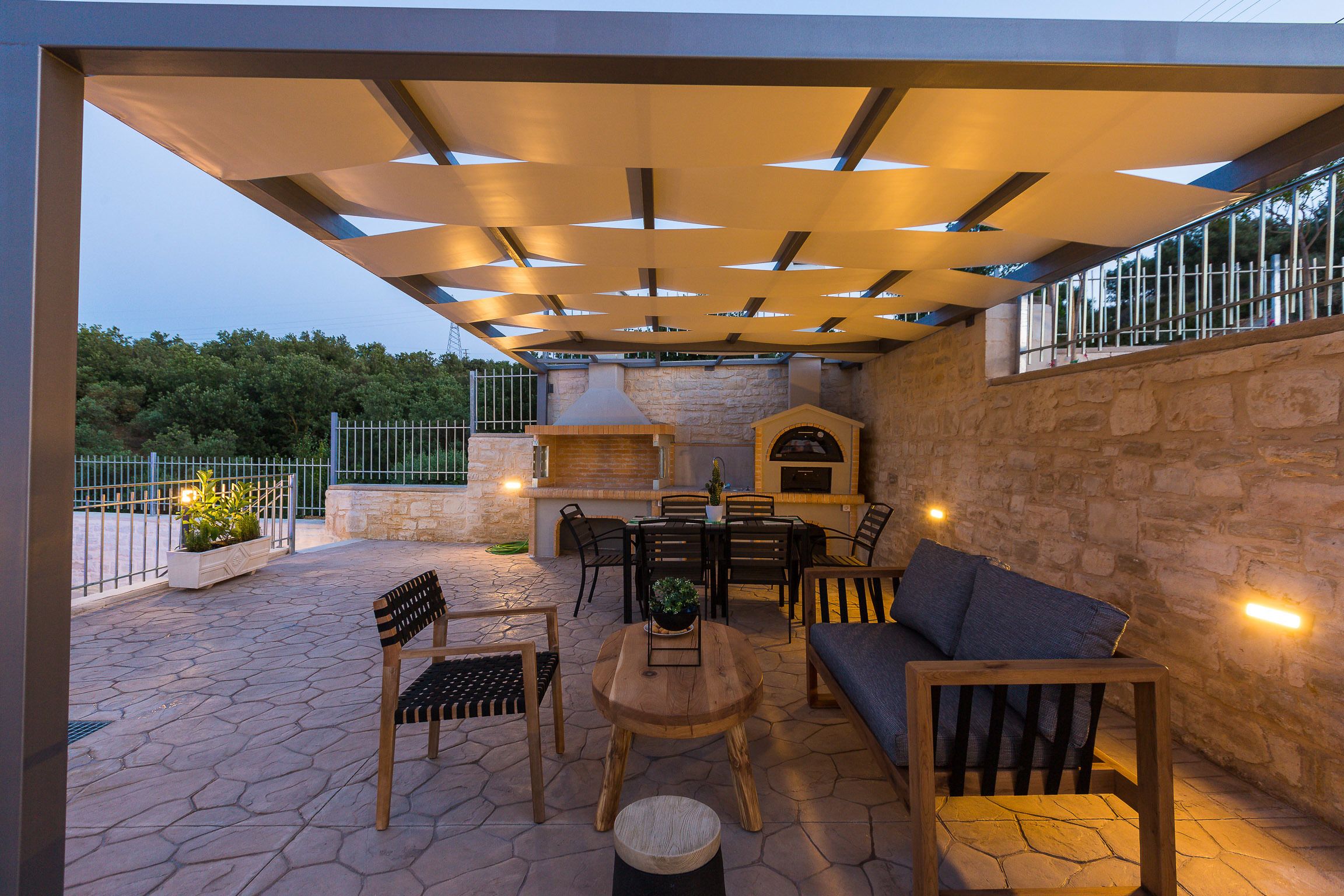 Un patio al aire libre con suelo de piedra, mobiliario de madera, una parrilla y un muro de piedra.